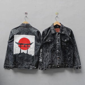 Può includere: Una giacca in denim nero lavaggio acido con un grande inserto quadrato bianco sul retro raffigurante un sole rosso e un portale torii nero. La parte anteriore presenta una chiusura a bottoni, due tasche sul petto e caratteri giapponesi bianchi.
