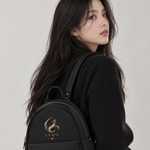 Puede incluir: Una mochila negra con un gráfico dorado de un zorro en forma de media luna sobre el texto YEON y caracteres coreanos. La mochila tiene una parte superior redondeada, un bolsillo frontal y cremalleras de color dorado en el compartimento.