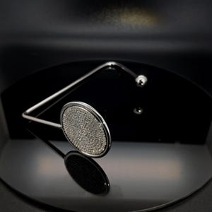Könnte beinhalten: Ein silberfarbener Taschenhaken aus Metall mit einer runden Scheibe an der Oberseite, die mit zahlreichen kleinen, klaren und funkelnden Steinen besetzt ist. Der Haken hat eine glatte, polierte Oberfläche und ein abgerundetes Ende.