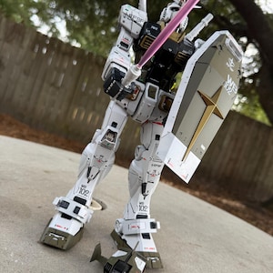 May include: A detailed white and olive green model robot figure with a shield and a pink energy sword. The robot has intricate panel lines and markings, with the number "102" visible on its legs. A small, olive green and black aircraft model is at its feet.