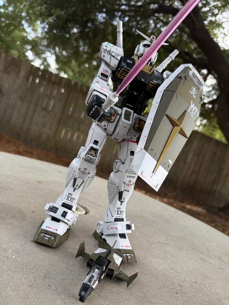 May include: A detailed white and olive green model robot figure with a shield and a pink energy sword. The robot has intricate panel lines and markings, with the number "102" visible on its legs. A small, olive green and black aircraft model is at its feet.