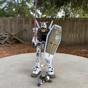 May include: A white, black, and gold robot action figure with a pink energy sword and a shield. The robot has the text "WB 102" on the shield and is standing on a concrete surface. A small, detached vehicle is at its feet.