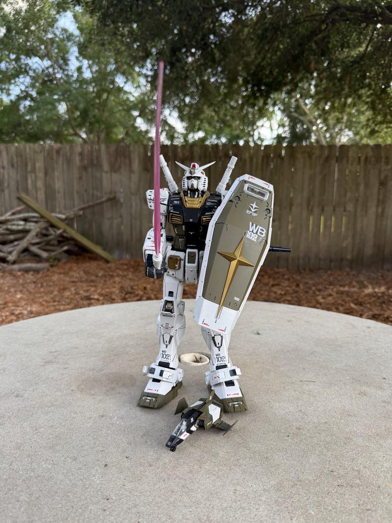 May include: A white, black, and gold robot action figure with a pink energy sword and a shield. The robot has the text "WB 102" on the shield and is standing on a concrete surface. A small, detached vehicle is at its feet.
