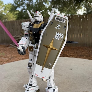 May include: A detailed model of a white, black, and gold robot with a shield and a pink energy sword. The robot has the text "EFSF" and "WB 102" on its body. The model is standing on a concrete surface.
