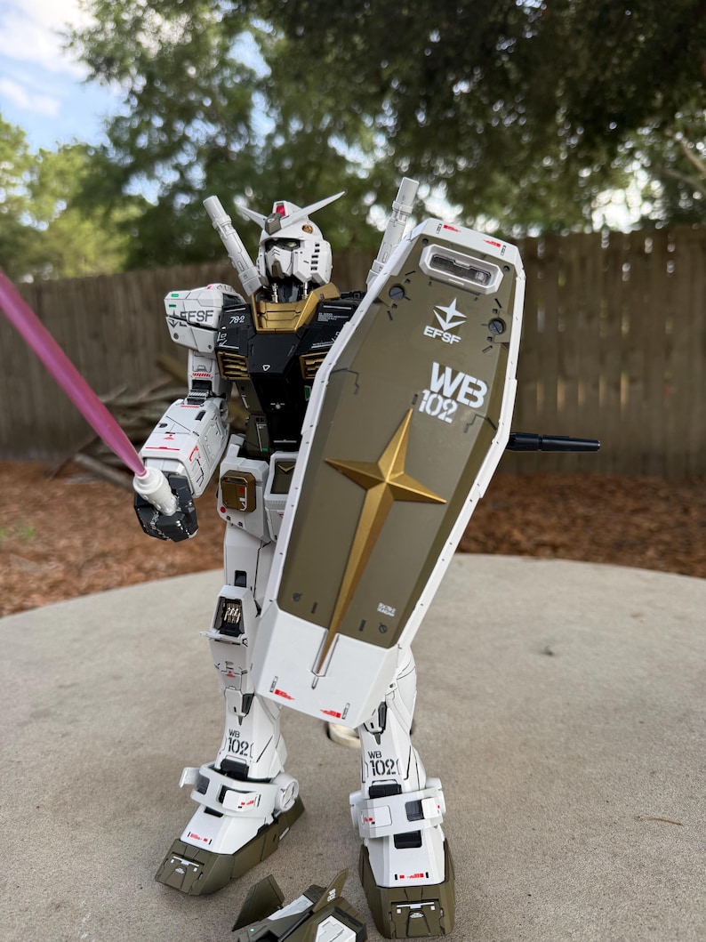 May include: A detailed model of a white, black, and gold robot with a shield and a pink energy sword. The robot has the text "EFSF" and "WB 102" on its body. The model is standing on a concrete surface.