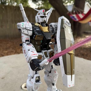 May include: A detailed model of a white, black, and gold robot with a shield and a pink energy sword. The robot has multiple markings and the text "EFSF" on its shoulder. The model is standing on a concrete surface.