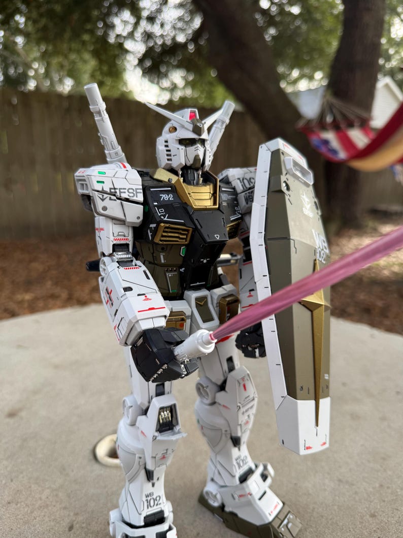 May include: A detailed model of a white, black, and gold robot with a shield and a pink energy sword. The robot has multiple markings and the text "EFSF" on its shoulder. The model is standing on a concrete surface.