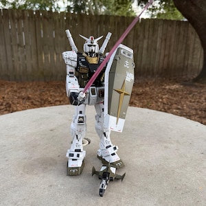 May include: A detailed model of a white, olive green, and gold robot with a red energy sword and shield. The robot has intricate mechanical details and is posed as if in action. The shield has a gold star and the letters "WB" on it.