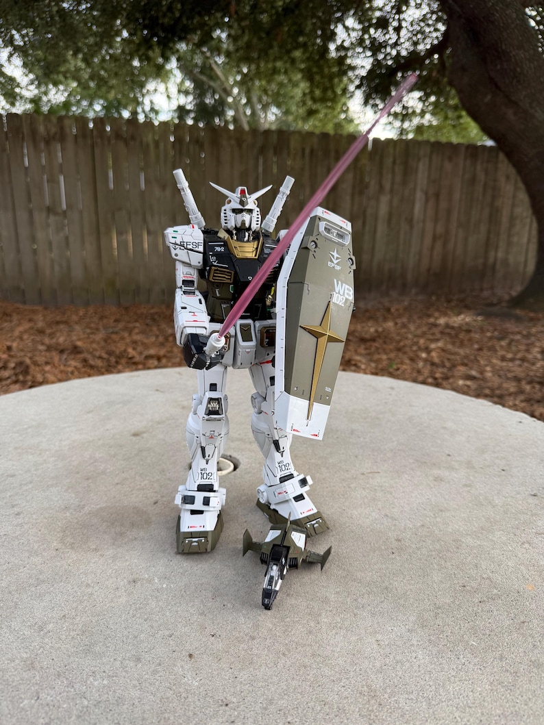 May include: A detailed model of a white, olive green, and gold robot with a red energy sword and shield. The robot has intricate mechanical details and is posed as if in action. The shield has a gold star and the letters "WB" on it.