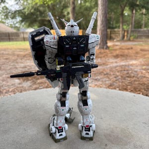 May include: A detailed white and black model robot with a shield and a rifle. The robot has red and green accents and is standing on a concrete surface. The background is a blurred view of trees and a house.