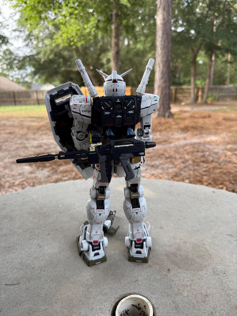 May include: A detailed white and black model robot with a shield and a rifle. The robot has red and green accents and is standing on a concrete surface. The background is a blurred view of trees and a house.
