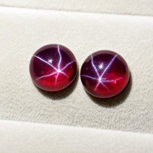 Pode incluir: Duas pedras preciosas cabochão redondas de cor vermelho profundo, cada uma apresentando um efeito de estrela de seis raios conhecido como asterismo. As pedras têm uma superfície lisa e polida que reflete a luz, criando um padrão de estrela branco.