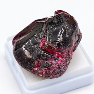 May include: A raw, unpolished gemstone with a deep, dark red and black coloration. The stone features a mix of smooth, reflective dark surfaces and rough, granular textured areas with vibrant magenta-red crystalline inclusions throughout.
