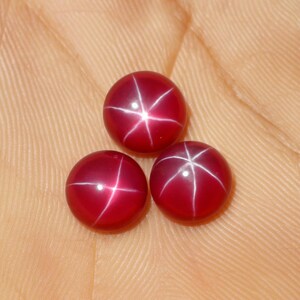 Può includere: Tre pietre preziose cabochon rotonde di colore rosso intenso, ognuna caratterizzata da un distinto motivo a stella a sei raggi noto come asterismo. Le pietre hanno una superficie bombata, liscia e lucida che riflette la luce creando un effetto a stella.