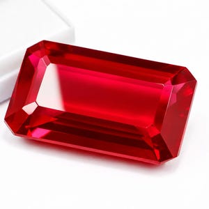 May include: A rectangular, faceted gemstone with a vibrant, deep red colour. The stone features a clear, polished surface with sharp, geometric edges and a brilliant, reflective quality, positioned next to a white, rectangular storage container.