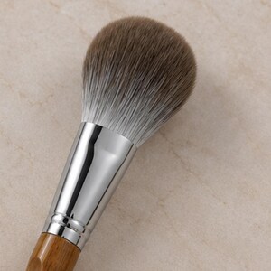 May include: A make-up brush featuring a rounded head of soft, dense bristles that transition from a dark brown top to a white base. The brush has a polished, reflective silver metal ferrule connecting the bristles to a smooth, light brown wooden handle.