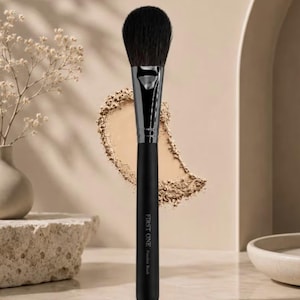May include: A makeup brush with a matte black handle, a silver ferrule, and soft black bristles. The handle features the text FIRST ONE Powder Brush printed in white. The brush is positioned vertically against a neutral background.