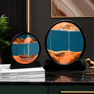 May include: Two circular sand art displays with black frames and stands, featuring orange sand and blue liquid. The sand creates mountain-like formations within the glass, with fine streams of sand falling through the blue space between the layers.