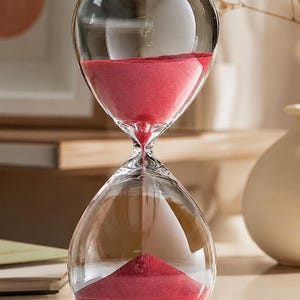May include: A clear glass hourglass featuring a rounded, bulbous shape with a narrow neck. The interior contains fine, vibrant red sand that is currently flowing from the top chamber into the bottom chamber, forming a small, conical pile.