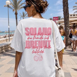 Könnte beinhalten: Weißes T-Shirt mit rosa Text auf dem Rücken. Der Text lautet "SOLANG ich noch Puls nach drei Promille hab BRENNT mein Herz für Malle lichterloh" mit zwei Bierkrügen darunter. Das T-Shirt wird im Freien getragen.