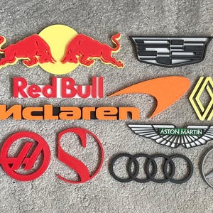 Könnte beinhalten: Eine Sammlung verschiedener Automobilmarken-Logos und Embleme, darunter Red Bull, McLaren, Ferrari, Cadillac, Renault, Aston Martin, Audi, Mercedes-Benz, Lotus, Alpine und Haas, ausgeführt als dreidimensionale Wand-Schilder.