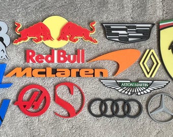May include: A collection of various automotive brand logos and emblems, including Red Bull, McLaren, Ferrari, Cadillac, Renault, Aston Martin, Audi, Mercedes-Benz, Lotus, Alpine, and Haas, rendered as three-dimensional wall-mounted signs.