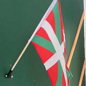 Pode incluir: Uma bandeira basca, conhecida como Ikurrina, com um fundo vermelho, uma cruz de Santo André verde e uma cruz branca sobreposta. A bandeira está presa a um mastro de madeira com um remate dourado no topo e montada na parede por um suporte preto.