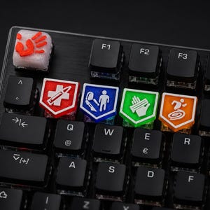 Könnte beinhalten: Eine mechanische Tastatur mit individuellen Artisan-Tastenkappen. Eine weiße Taste zeigt einen roten Handabdruck. Vier schildförmige Tasten in Rot, Blau, Grün und Orange tragen weiße Symbole, die eine Spritze, ein Messer, Verbände und eine laufende Figur darstellen.