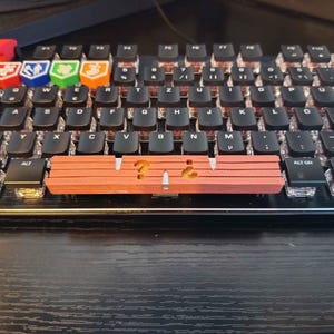 Könnte beinhalten: Eine schwarze mechanische Tastatur mit individuellen Tastenkappen, darunter eine lange terrakottafarbene Leertaste mit ausgeschnittenem Fragezeichen und umgekehrtem Fragezeichen sowie mehrere bunte quadratische Tastenkappen mit verschiedenen Symbolen.