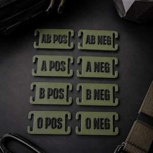 Könnte beinhalten: Ein Set aus acht rechteckigen, olivgrünen Blutgruppen-Aufnähern mit schwarzer Prägeschrift. Die Aufnäher sind mit AB POS, AB NEG, A POS, A NEG, B POS, B NEG, O POS und O NEG beschriftet. Jeder Aufnäher verfügt über integrierte seitliche Schlitze zur Befestigung.