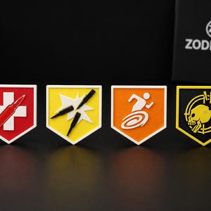 Könnte beinhalten: Vier schildförmige Aufnäher mit verschiedenen grafischen Motiven: ein roter Aufnäher mit weißem Kreuz und Patrone, ein gelber mit weißem Stern und drei Patronen, ein orangefarbener mit weißer laufender Figur und ein schwarzer mit gelbem Totenkopf.