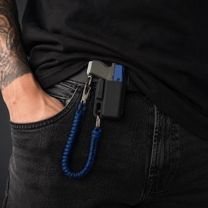 Könnte beinhalten: Eine schwarze Halterung mit einem blauen Schlüsselband, das an einer Gürtelschlaufe befestigt ist. Die Halterung hält ein kleines, grau-blaues Objekt. Die Person trägt ein schwarzes T-Shirt und eine dunkelblaue Jeans. Ein Tattoo ist auf dem Arm der Person sichtbar.