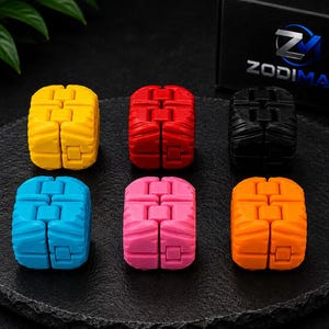 Könnte beinhalten: Sechs Fidget-Cubes in Gelb, Rot, Schwarz, Blau, Pink und Orange, angeordnet in zwei Reihen. Jeder Würfel hat eine segmentierte, strukturierte Oberfläche mit sichtbaren Scharnieren. Eine schwarze Box mit dem weißen und blauen Text ZODIMAK ist zu sehen.