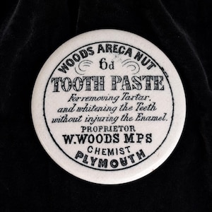May include: A round white ceramic lid featuring black printed text that reads: WOODS ARECA NUT 6d TOOTH PASTE For removing Tartar, and whitening the Teeth without injuring the Enamel. PROPRIETOR W.WOODS MPS CHEMIST PLYMOUTH. The lid has a smooth finish.