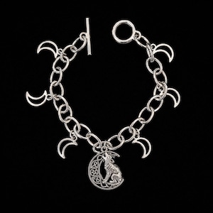 Op de afbeelding: Een zilverkleurige schakelarmband met een staafsluiting en meerdere maansikkelbedels. Een centrale hanger toont een wolf die naar een maansikkel huilt, versierd met kleine stervormige uitsparingen en ingewikkelde knoopmotieven.