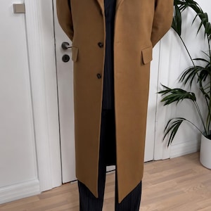 May include: A long camel-coloured overcoat with notched lapels and two front buttons, worn over a dark charcoal pinstriped suit jacket, a white collared shirt, a dark brown tie, and dark charcoal pinstriped trousers with black leather shoes.