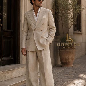 May include: A beige double-breasted suit with a white shirt and a pocket square. The suit features wide-leg trousers and a structured blazer with buttons. The text ELITESTUDIO ATTIRE USA PREMIUM TAILORED TIMELESS is visible on the right side.