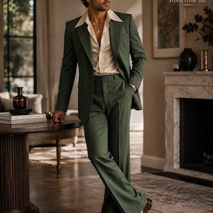 Op de afbeelding: Een donker olijfgroen pak bestaande uit een blazer met brede revers en een bijpassende broek met wijde pijpen. Het pak wordt gecombineerd met een lichtbeige overhemd met een open kraag. De tekst ELITE STUDIO ATTIRE USA staat rechtsboven.
