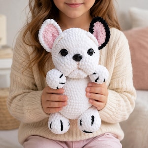 Può includere: Un cane di peluche all'uncinetto bianco con una finitura in filato testurizzato, caratterizzato da grandi occhi neri, un naso nero e due orecchie dritte. Un orecchio è bianco con l'interno rosa, mentre l'altro è nero con l'interno rosa.