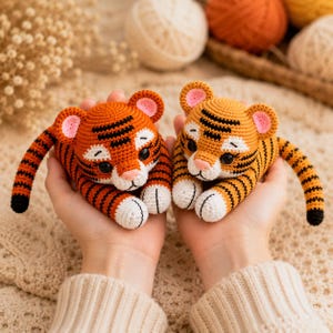 Puede incluir: Dos pequeños juguetes de tigre tejidos a ganchillo con cuerpos de hilo naranja y amarillo, rayas negras, patas blancas y orejas internas rosas. Cada tigre presenta ojos oscuros redondos, una pequeña nariz rosa y una larga cola rayada.