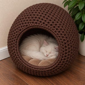 Pode incluir: Uma cama esférica para animais de estimação em crochê castanho escuro com uma abertura circular. Dentro da cama, um gato branco de pelo comprido está enrolado e a dormir sobre uma almofada macia e tufada em castanho claro.