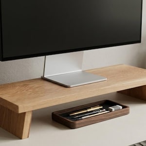 May include: A light-coloured wooden monitor stand with a rectangular top and angled legs, supporting a computer screen. Beneath the stand sits a dark wooden rectangular tray containing two pens, a mechanical pencil, and a small rectangular eraser.