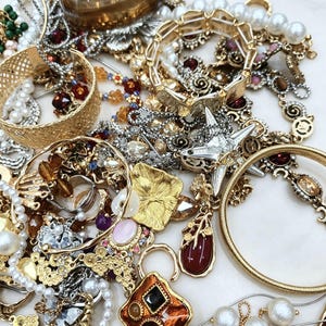 May include: A collection of assorted jewelry including gold-tone metal bangles, a brown translucent cuff, strands of white faux pearls, and various pendants featuring red, amber, and black stones. The assortment includes textured metal links and chains.