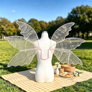 May include: A pair of translucent fairy wings with a delicate gold glitter vein pattern, attached to a light pink frame. The wings are mounted on a white mannequin torso, showcasing the intricate, shimmering design of the sheer fabric material.