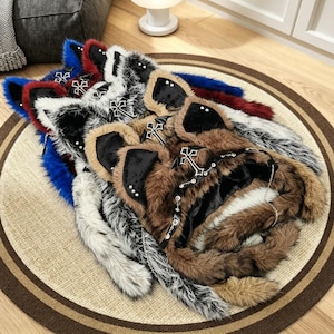 May include: A collection of faux fur animal ear hats in brown, white, black, red, and blue. Each hat features plush ears, a silver-toned cross emblem, metal studs, and a thin chain strap. The hats are layered, showcasing varied fur textures.
