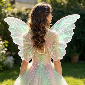 May include: A pair of large, iridescent fairy wings with a translucent, shimmering finish and delicate vein detailing. The wings are attached to a pastel-coloured dress featuring a corset-style back with white pearl-like bead trim and lacing.