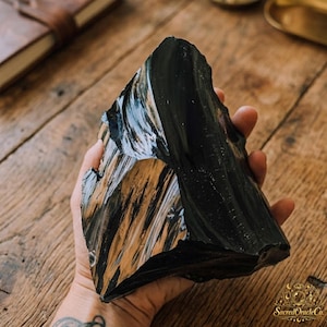 Puede incluir: Un trozo grande y dentado de obsidiana negra con una superficie brillante y reflectante. La piedra presenta bordes afilados e irregulares y una textura suave similar al vidrio con variaciones sutiles en el reflejo de la luz sobre su forma volcánica.