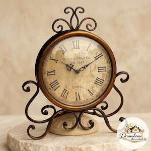 Puede incluir: Un reloj de mesa redondo con marco de metal color bronce y base con volutas ornamentadas. La esfera presenta un fondo beige desgastado con números romanos negros, manecillas negras y el texto Winston Clock Co. Est. 1845 y Quartz.