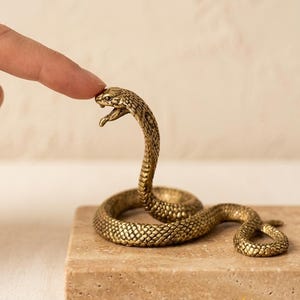 Op de afbeelding: Een messingkleurig metalen slangenbeeldje met een textuur van schubben en een open mond, geplaatst op een vierkante, lichtbeige stenen voet. De slang is opgerold met de kop omhoog, voorzien van gedetailleerde ogen en een zichtbare tong.