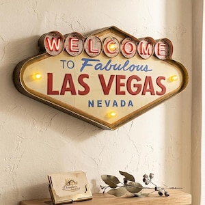 Könnte beinhalten: Ein Schild im Vintage-Stil von Las Vegas mit den Worten "Welcome to Fabulous Las Vegas Nevada" in Rot und Blau. Das Schild hat einen goldfarbenen Rahmen und ist mit kleinen Lichtern beleuchtet. Unterhalb befindet sich ein Holzregal mit einem Kartenhalter und einem dekorativen Zweig.